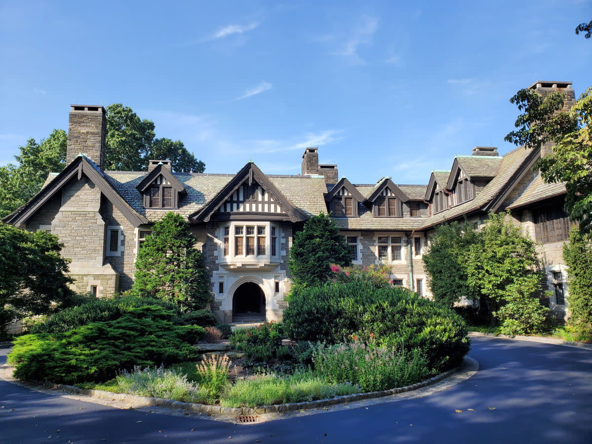 View of the primary northeast façade of the house, facing southwest, along the entrance drive. The house is positioned within an Olmsted landscape featuring mature trees and formal gardens, creating a private estate setting with the city of Philadelphia.